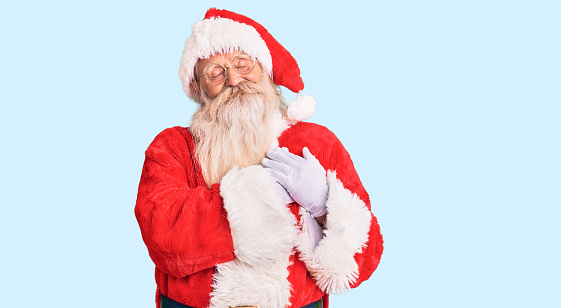 Old senior man with grey hair and long beard wearing traditional santa claus costume smiling with hands on chest with closed eyes and grateful gesture on face. health concept.