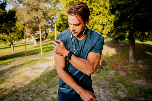 Injured young caucasian male athlete looking concerned holding shoulder with pain after exercising outdoors in the sun with smartwatch for tracking activities and ear phones for listening to music. High quality photo