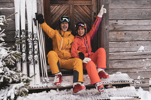 cheerful very happy mature adult sporty ski couple sitting at wooden mountain hut smiling laughing at camera