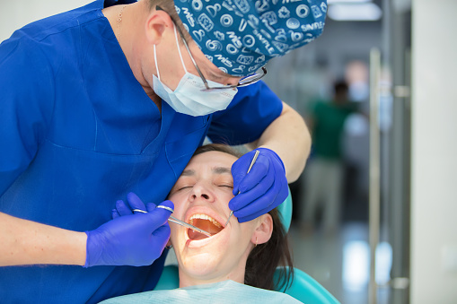 Dentist young woman treats a patient a man. The doctor uses disposable gloves, a mask and a hat. The dentist works in the patient's mouth, uses a professional tool