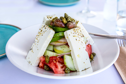 White bowl of Greek Salad with feta cheese, capers, cucumber, tomatoes, peppers, olives, olive oil drizzled with spoon behind the salad, on white table cloth, green aqua side plates.