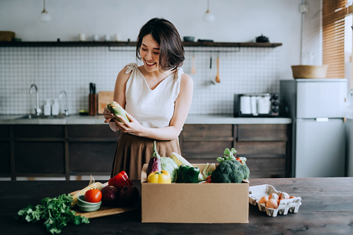 Beautiful smiling young Asian woman received a full box of colourful and fresh organic groceries ordered online by home doorstep delivery service. She is sorting out the groceries and preparing to cook a healthy meal