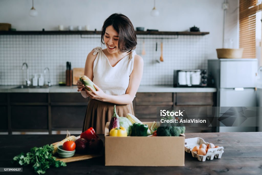 Beautiful smiling young Asian woman received a full box of colourful and fresh organic groceries ordered online by home doorstep delivery service. She is sorting out the groceries and preparing to cook a healthy meal Supermarket Stock Photo