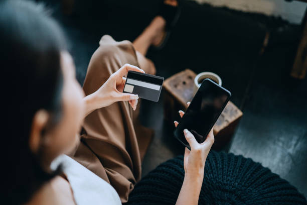 sur la vue d’épaule de la jeune femme asiatique se refroidissant à la maison, s’asseyant sur le plancher dans la chambre à coucher, appréciant une tasse de café et faisant des achats en ligne avec le smartphone tout en faisant le paiement mobile a - hotel wireless technology bedroom hotel room photos et images de collection