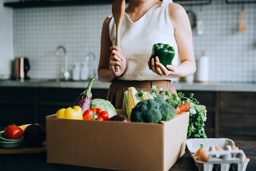Young Asian woman received a box full of colourful and fresh organic groceries ordered online. She is planning to cook a healthy meal in the kitchen at home