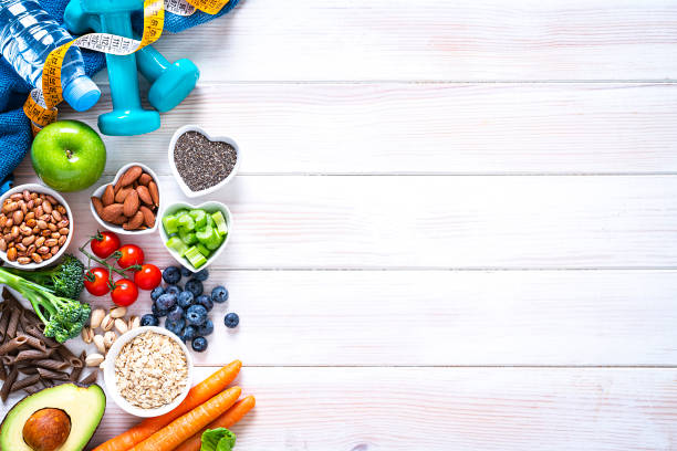 Healthy eating and exercising concepts: overhead view of exercising items and healthy food to keep a healthy lifestyle. The composition arranged side by side at the left border of a white table leaving useful copy space and includes dumbbells, blue towel, tape measure, a water bottle and some fruits, vegetables and nuts like broccoli, almonds, avocado, lettuce, carrots, oat, beans, tomato, blueberries, a green apple, whole grain pasta, pistachio, chia seeds and celery. High resolution 42Mp studio digital capture taken with SONY A7rII and Zeiss Batis 40mm F2.0 CF lens