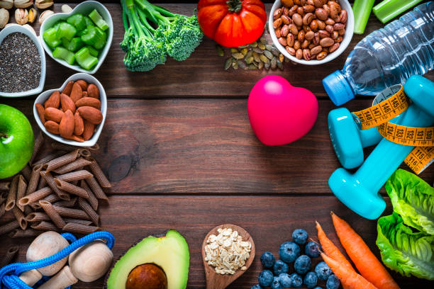 Sports and healthy food frame: fruits, vegetables, nuts, dumbbels and tape measure. Copy space at the center Healthy eating and exercising concepts: overhead view of exercising items and healthy food to keep a healthy lifestyle. The composition arranged side by side all around the border of a wooden table leaving useful copy space and includes dumbbells, tape measure, a water bottle and some fruits, vegetables and nuts like celery, broccoli, almonds, pistachio, whole grain pasta, avocado, lettuce, carrots, oat, beans, tomato, blueberries, a green apple, chia seeds and pumping seeds. High resolution 42Mp studio digital capture taken with Sony A7rII and Sony FE 90mm f2.8 macro G OSS lens celery heart stock pictures, royalty-free photos & images