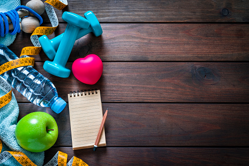 Healthy lifestyle and fitness background: top view of dumbbells, jump rope, water bottle, tape measure, towel, green apple, note pad and pencil arranged at the left of a brown wood plank leaving useful copy space for text and/or logo at the right. A red heart is included in the composition. Predominant colors are blue and brown. High resolution 42Mp studio digital capture taken with SONY A7rII and Zeiss Batis 40mm F2.0 CF lens