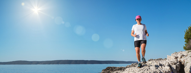 Middle-aged female dressed running sporty clothes and pink cap enjoying morning jogging along the rocky calm sea coast. Sporty people trail running activities on vacation time concept image.