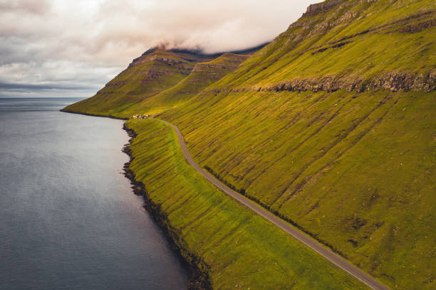 Faroe Islands Green Coastal Road Kunoy Island Sunset Light Faroe Islands Kunoy Island empty coastal road along the grass covered fjord hills. Aerial Drone Point of View towards the cloud covered mountain peaks and North Atlantic Ocean horizon. Kunoy Island, Faroe Islands, Kingdom of Denmark, Nordic Countries, Europe eysturoy stock pictures, royalty-free photos & images