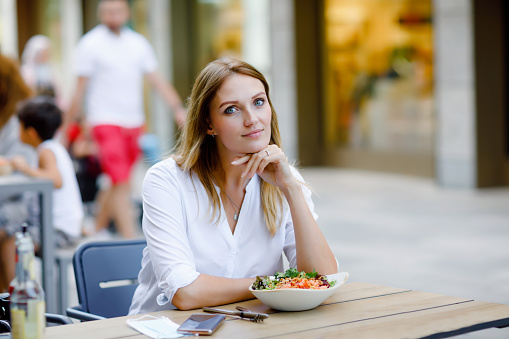 Young beautiful woman eating fresh salad in outdoor restaurant. Woman enjoying lunch bowl with fresh vegetables. Healthy food. On summer day