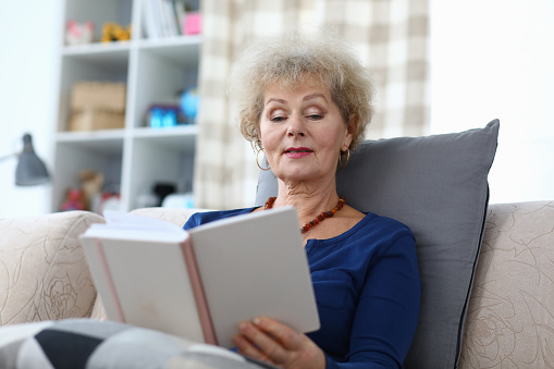 Grandmother sits at home and read with enthusiasm. Self-isolation book an elderly woman at home. An elderly woman is reading book at home. Affordable entertainment for adult parents