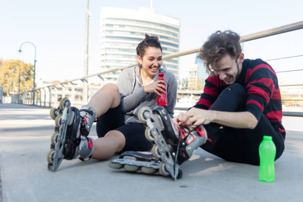 A happy young couple is drinking water and cheerfully talking while tying their roller skates at a waterfront park