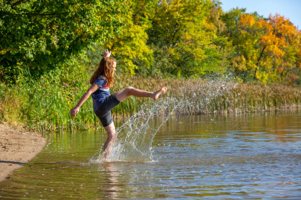 Girl standing in shallow water kicking the water to make it splash up. The girl has curly copper colored hair. Taken on an early autumn evening with the trees just starting to change colors.
