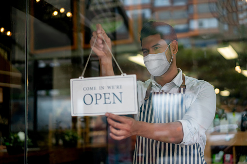 Portrait of a Latin American man working at a grocery store wearing a facemask and hanging an open sign on the door â reopening of businesses concepts