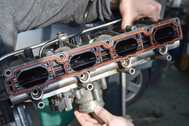 A car mechanic holds an intake manifold from a car engine A car mechanic in a gray work suit holds an exhaust manifold with a throttle removed from an automobile internal combustion engine throttle photos stock pictures, royalty-free photos & images