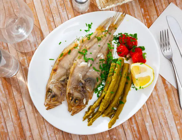 Whole oven baked rooster fish with marinated asparagus and tomatoes on white plate