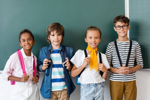 istock Multicultural schoolchildren with backpacks looking at camera near chalkboard 1274064252