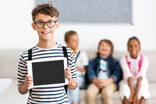 Selective focus of schoolboy in eyeglasses showing digital tablet with blank screen in classroom