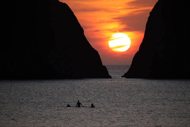 Three silhouetted unidentifiable surfers return from the sea in front of Gull Rocks at Holywell Bay in Cornwall as the sun sets behind the clouds and the V shape formed by the natural rock formations.