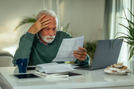 Senior man at home going over paperwork and using a laptop