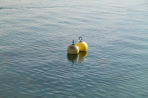 two water buoys floating in the water one white the other yellow plastic remains hang on the yellow buoy during the day in good weather without people