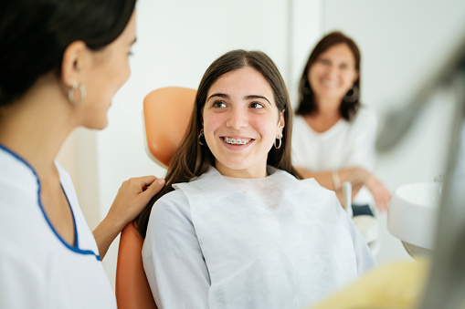 Hispanic teenage girl with braces sitting in dentist chair and smiling at young female orthodontist after an encouraging examination.