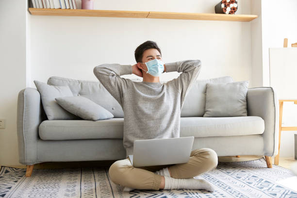 young asian man wearing mask sitting on floor at home young asian business man staying at home wearing mask sitting on carpet working using laptop computer stay at home order stock pictures, royalty-free photos & images