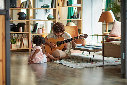 Mother in mid 20s sitting on floor of family living room with 3 year old daughter and playing acoustic guitar.