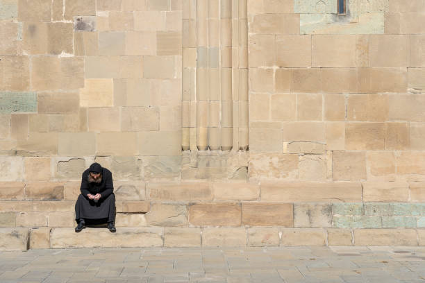 lonely old monk in black clothes sits on stairs near the old orthodox cathedral in town mtskheta near tbilisi, georgia - mtskheta imagens e fotografias de stock