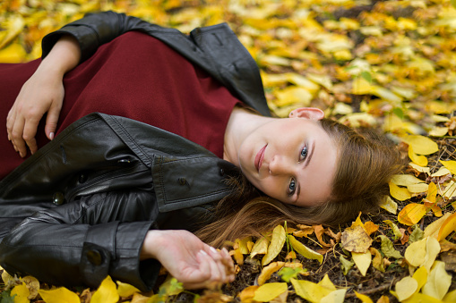 A beautiful blue-eyed girl in a red dress and a black leather jacket is lying on the ground on the yellowed leaves. Happy young woman in park on sunny autumn day.  Girl in the autumn forest. Autumn