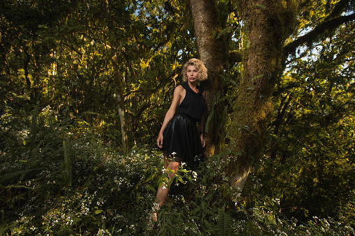 Beautiful blonde in a black dress with a hood stands near a tree in the forest. The girl got lost in the woods brooding dreamy Young women walking through the woods. Fashion style High quality photo