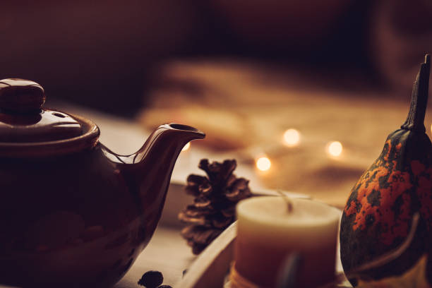 Close up, cut out of a vintage tea kettle placed in the wooden tray with a pine cone placed next to it, a candle and decorative orange and green pumpkin.