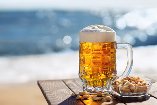 mug of cold beer and salty peanuts in bowl on table with sea on the background