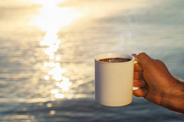 male hand with mug of hot coffee against sea at sunrise as background