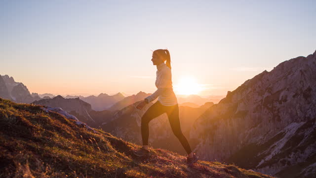 Fit woman athlete maintaining a healthy lifestyle, walking on the edge in mountain terrain over rocky trails and grassy slopes, illuminated by sunset light