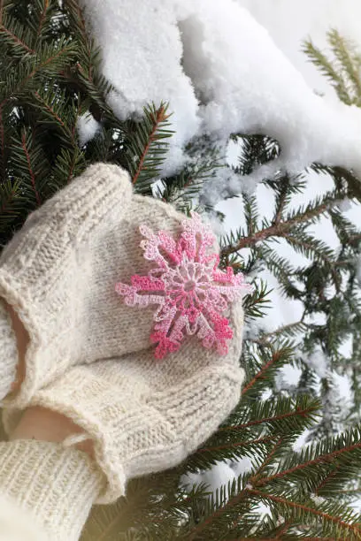 pink knitted snowflake in the hands of dressed in mittens against the background of snow-covered fir branches