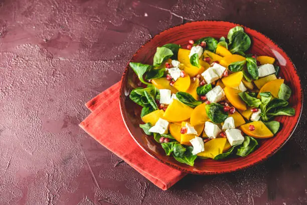 Healthy salad with persimmon, doucette (lambs-lettuce, cornsalad, feld salad) and feta cheese on a red plate on a red background. Superfoods Vitamin autumn or winter persimmon salad