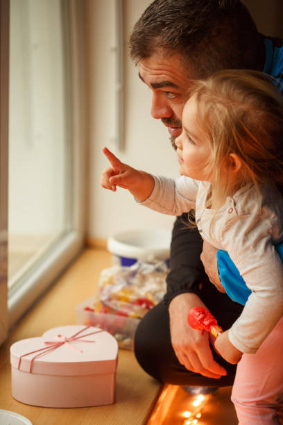 Side view of affectionate father holding his cute little daughter while they are looking outside, through the window and she is pointing at something.