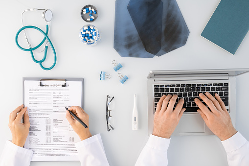 Flatlay of hands of two clinicians making medical notes and typing by laptop surrounded by x-ray images, pills, stethoscope and others