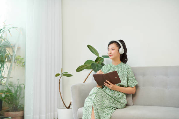 istock mage of focused asian woman using headphones and writing down notes while sitting on sofa at home 1274870812