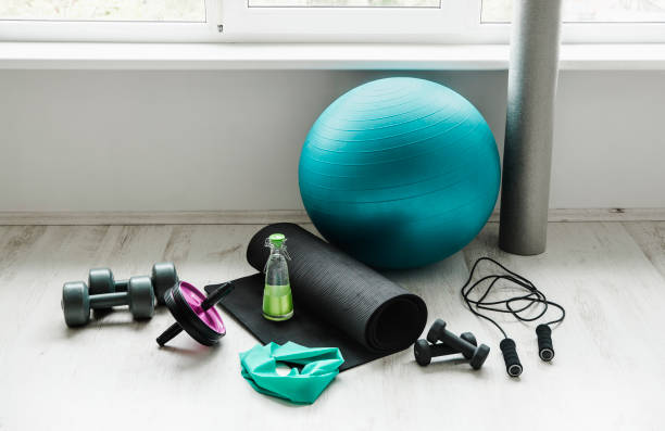 Group of different exercising equipment on white home gym floor. Fitness ball, round foam roller, resistance exercise latex band, jumping rope, dumbbells, yoga mat. Fit lifestyle concept. stock photo