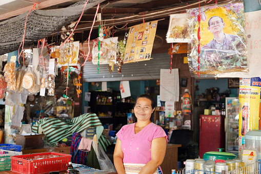 Rural thai village vendor woman at her shop  - woman is standing in opened shop and is looking. Around woman are many things and packeged food for selling, Above woman are many pictures of and with life of King Bhumipol Rama IX of Thailand