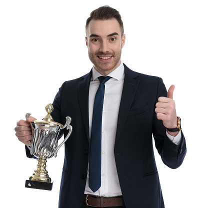 Positive businessman holding trophy and gesturing ok while standing on white studio background