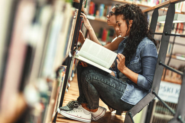 istock Two female students reads book by the book shelf at the library. 1274987747