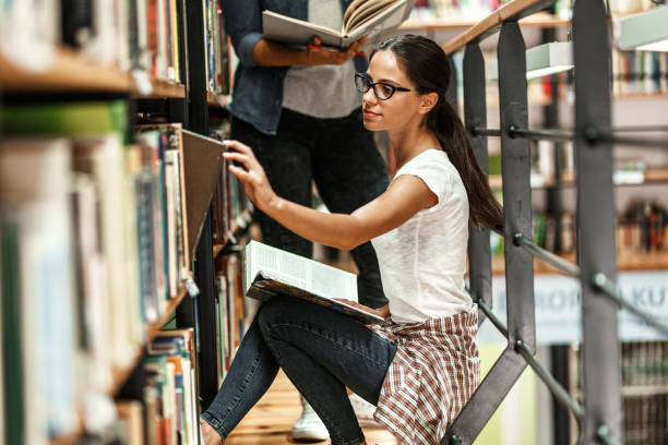 istock Young female student standing by the book shelf at the library and reading a book. 1274987949