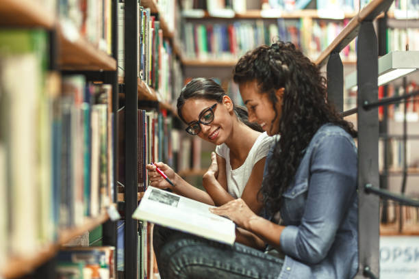 istock Two female students reads book by the book shelf at the library. 1274988355