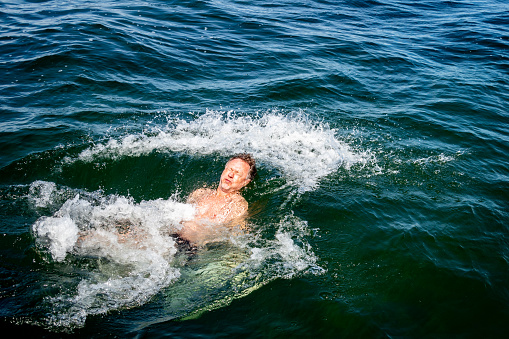 Caucasian male with painful facial expression jumps backwards into icy splashing water in the ocean of the Baltic Sea. Swim in the cold freshwater surrounding the island Gotland at the city Visby.