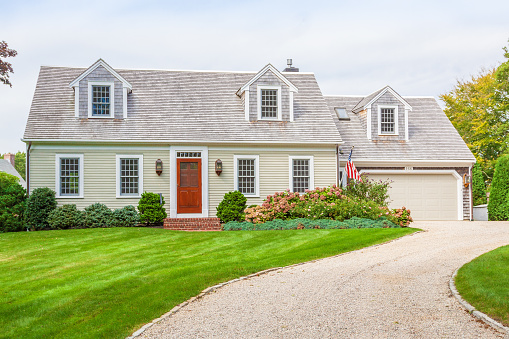 New England House with Beige Clapboard Exterior, Grey Shingle Roof, Gravel Driveway, Garage, Red Door, American Flag and Landscaped front yard with green grass, bushes and flowers. The House is in Chatham, Cape Cod, Massachusetts. Canon EF 24-105mm/4L IS USM Lens.