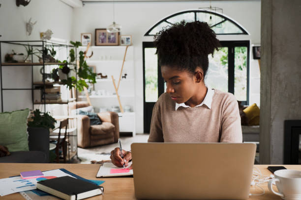 istock Mixed race female student studying online writing notes on notepad reading off laptop sitting in modern kitchen 1275047751